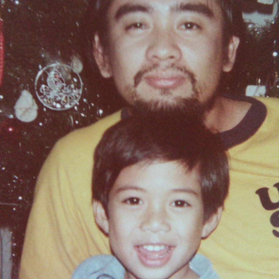 A probably mid to late 30s Filipino Father with a beard and mustache poses in front of a Christmas tree with his son, the author of this post. They have a striking resemblance.